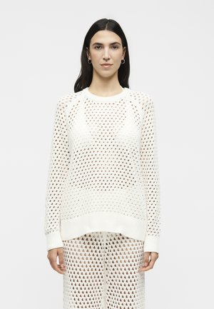 Γυναίκα με μακριά σκούρα μαλλιά φοράει λευκό πλεκτό πουλόβερ και παντελόνι ταιριαστό, μπροστά σε λευκό φωτεινό φόντο.