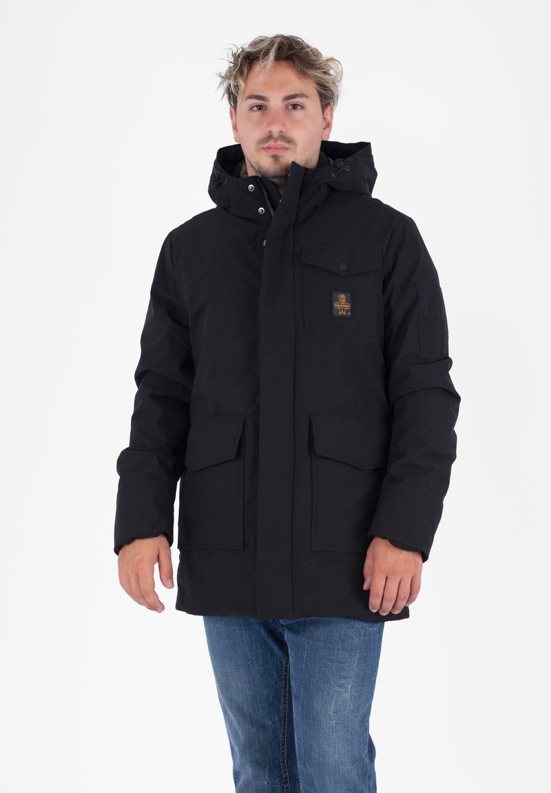 Hombre que lleva una chaqueta de invierno con capucha negra y bolsillos delanteros, y unos jeans azules, de pie frente a un fondo blanco.