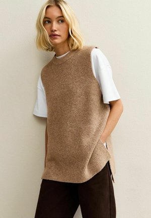 Brauner, gerippter Strick-Ärmelloser Pullover über einem weißen T-Shirt, mit seitlichen Schlitzen und lockerem Schnitt, vor neutralem Hintergrund.