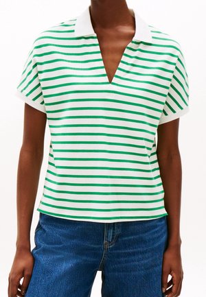 Persoon in een wit shirt met groene horizontale strepen, korte mouwen, een witte kraag en blauwe jeans tegen een effen achtergrond.