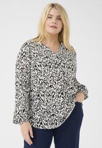 Langärmlige Bluse mit schwarz-weißem Muster und V-Ausschnitt, zwei Brusttaschen und lockerer Passform, kombiniert mit dunklen Jeans.