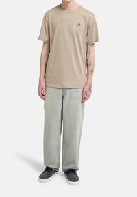 Μπεζ βαμβακερό μπλουζάκι με στρογγυλή λαιμόκοψη, σετ με ανοιχτό πράσινο φαρδύ τζιν και μαύρα αθλητικά παπούτσια. Το μπλουζάκι έχει μικρό κεντημένο λογότυπο.