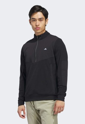 Jeune homme portant un haut noir à manches longues avec fermeture éclair au quart et pantalon beige clair, debout devant un fond clair uni.