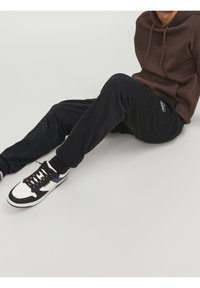 Černé tepláky s nášivkou loga, doplněné o hnědou mikinu s kapucí a bílé tenisky s černými a námořnickými akcenty. Látka vypadá měkká a strukturovaná.