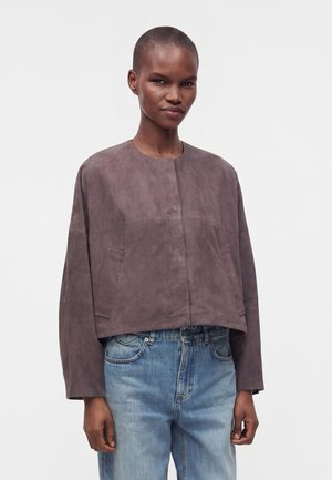 Person trägt eine lockere, kurz geschnittene braune Jacke mit langen Ärmeln und hellblauen Jeans, steht vor einem schlichten weißen Hintergrund.