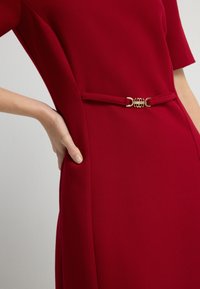 Rode jurk met een aansluitende silhouet en korte mouwen. Heeft een getailleerde taille met een gouden hardware riemdetail voor extra accent.