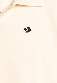 Camisa polo de suave tela beige con un logotipo negro bordado que presenta un diseño de estrella y flecha en el área izquierda del pecho.