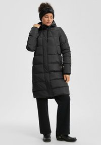 Sieviete, ģērbusies garā melnā piepūšamā mētelī ar kapuci, melnās biksēs, zābakos un melnā galvas lentes, stāv pretī neizteiksmīgai fona.