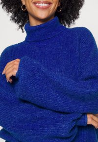Maglione blu a collo alto realizzato in tessuto a maglia testurizzato, con maniche ampie e una vestibilità rilassata. Primo piano che mette in evidenza il colletto e i polsini a coste.