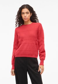Vrouw met krullend haar, gekleed in een rood gebreide textuursweater en zwarte broek, staande tegen een effen witte achtergrond.