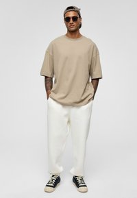 Beige oversized t-shirt met korte mouwen, witte sweatpants en zwarte sneakers met witte veters, alles tegen een lichtgrijze achtergrond.