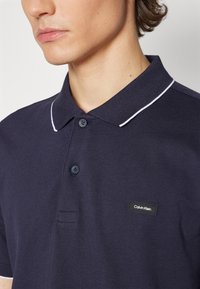 Polo de color azul marino con tejido texturizado, dos botones, cuello con ribete blanco y un parche del logo de Calvin Klein en negro.
