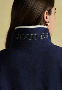 Námornícky fleecový kabát so bielym golierom, na ktorom je vyšitý text "JOULES" zlatou farbou. Hladká textúra, ležérny dizajn.