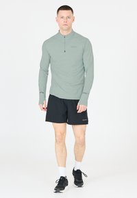 Lange mouwen, lichtgroene sporttop met een ritskraag; gecombineerd met zwarte shorts. Zwarte sneakers en witte sokken maken de outfit compleet.