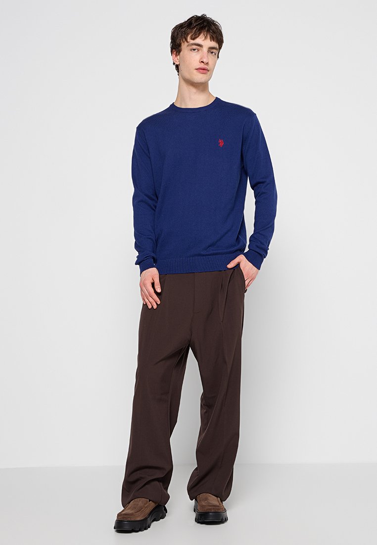 Marineblauer Strickpullover mit Rundhalsausschnitt und geripptem Saum, kombiniert mit locker sitzenden braunen Hosen und braunen Schuhen. Einfaches rotes Logodetail sichtbar.