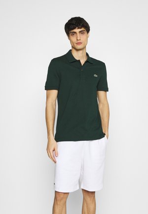Mörkgrön polotröja med krage och korta ärmar, prydd med en liten logotyp på vänster bröst, i kombination med vita shorts.