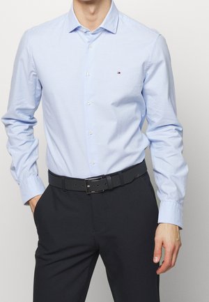 Homme portant une chemise habillée bleu clair boutonnée, rentrée dans un pantalon noir avec ceinture noire, debout avec une main dans la poche.