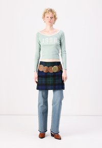 Crop top lung cu maneci lungi de culoare verde menta cu text alb, fustă de tip kilt cu carouri în tonuri de albastru și verde, curea maro cu accente circulare și blugi evazați de culoare albastră deschisă.
