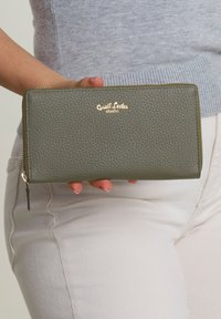 Portafoglio in pelle verde testurizzata con logo in tono oro, chiusura con zip e forma rettangolare. Tenuto in una mano, con uno sfondo neutro.
