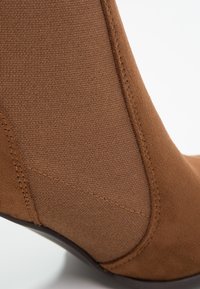 Gros plan sur une botte cheville en daim marron avec panneau latéral élastique texturé et coutures visibles.