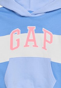 Ανοιχτό μπλε φούτερ με λευκή οριζόντια ρίγα. Περιέχει το λογότυπο "GAP" σε ροζ, φτιαγμένο από μαλακό υλικό με μπροστινή τσέπη.