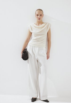 Donna che indossa un top smanicato crema con texture, pantaloni bianchi a gamba larga, scarpe nere appuntite, che tiene una pochette nera su sfondo bianco.