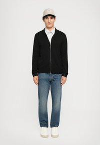 Schwarze Zip-Up-Sweatjacke, darunter ein weißes Hemd mit Kragen, blaue Jeans und weiße Sneaker, ergänzt durch eine hellfarbige Mütze.