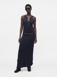 Femme portant une robe maxi noire sans manches avec une ouverture en forme de goutte sur le devant, ceinture cloutée, bottes noires et lunettes de soleil foncées, debout sur fond blanc.