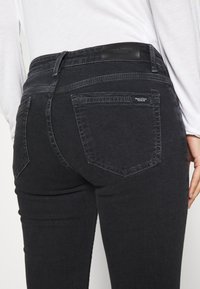 Jeans de mezclilla negra con un diseño ajustado. Sus características incluyen dos bolsillos traseros y un parche de cuero en la cintura. Textura suave y lisa.
