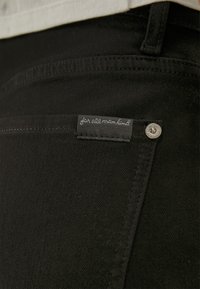 Jeans de mezclilla negra con un bolsillo visible, que presentan una etiqueta cosida que dice "para toda la humanidad" y un acento de botón plateado.