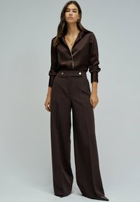 Mujer vestida con una camisa de satén marrón oscuro abotonada, pantalones de pierna ancha de talle alto, aretes dorados en forma de aro y collares dorados en capas, de pie contra un fondo gris.