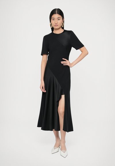 Fekete ruha rövid ujjakkal, aszimmetrikus kialakítással és oldalsó hasítékkal. Sima anyagból készült, áramló szoknyával. Fehér cipővel kiemelve.