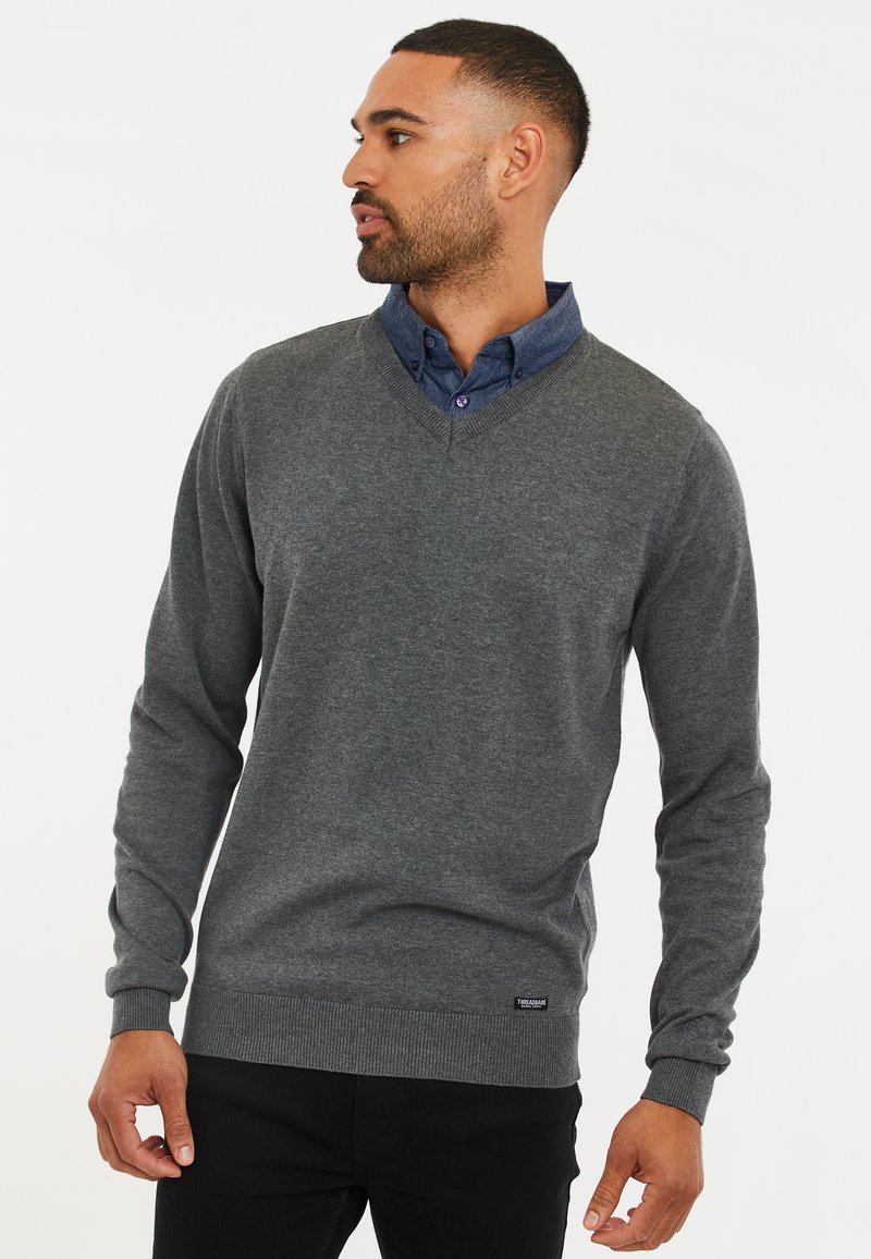 Maglione grigio a V con maniche lunghe, texture morbida, polsini e orlo a costine. Indossato sopra una camicia blu con colletto e bottoni.