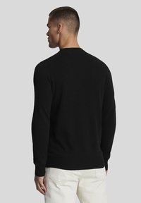 Fekete pulóver hosszú ujjakkal és kerek nyakkivágással, sima anyagból készült. A hátoldal egyszerű mintázatú, világos színű nadrággal kombinálva.