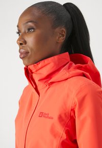 Veste imperméable orange vif avec un col montant et une capuche. Présente un logo sur la poitrine et une texture lisse et mate.