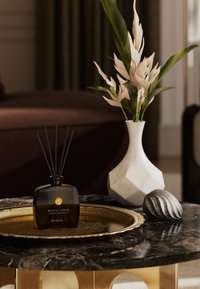 Sort glasdiffuserflaske mærket "MYSTIC CEDAR" med sorte rør, på et guldfad ovenpå et mørkt marmorbord, ved siden af en hvid geometrisk vase og en struktureret sten.