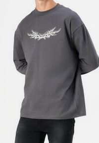 Homme portant un T-shirt à manches longues gris foncé avec un motif tribal abstrait blanc sur la poitrine et un pantalon noir, debout avec les mains derrière le dos.