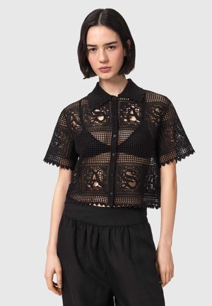 Młoda kobieta z krótkimi ciemnymi włosami, ubrana w czarny szydełkowany top zapinany na guziki na czarnym biustonoszu oraz luźne czarne spodnie na jednolitym tle.