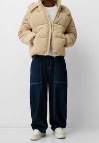 Beige corduroy puffer jacket with a high collar, zip closure, and plush texture, paired with dark denim wide-leg pants and beige sneakers.
