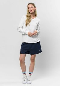 Biely dlhý rukáv top s výstrihom do V a malým logom, v kombinácii s námorníckymi šortkami. Model má na sebe pruhované ponožky a biele tenisky. Hladká textúra.