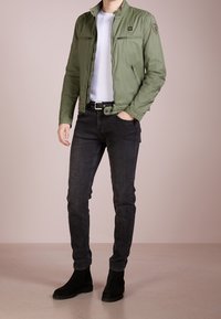 Veste légère verte avec fermeture éclair à l'avant, deux poches latérales et une poche poitrine. Portée sur une chemise blanche, associée à un jean noir et des bottes.
