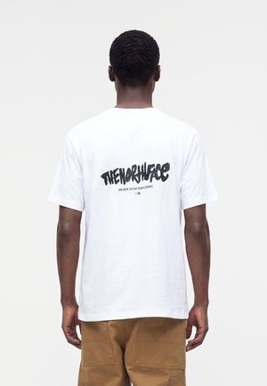 Homme portant un t-shirt blanc avec un texte noir "The North Face Never Stop Exploring" dans le dos et un pantalon marron, debout devant un fond uni.