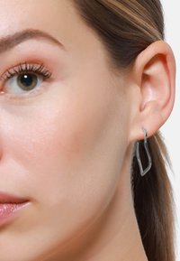 Silberne geometrische Ohrringe mit einem eleganten, offenen Umrissdesign. Die metallische Oberfläche reflektiert das Licht, und sie werden an einem Ohr getragen.