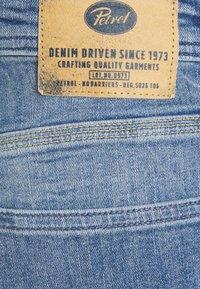 Denimtyg med en ljusblå nyans, med en läderetikett där det står "Petrol" och sömmar i gult. Texturerad yta med blekta inslag.