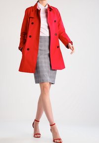 Czerwony trencz z czarnymi guzikami, noszony na szarej spódnicy ołówkowej w kratę. W zestawie z czerwonymi sandałami na wysokim obcasie. Gładka tkanina i dopasowany krój.