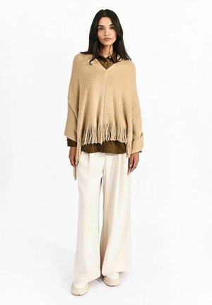 Donna che indossa un poncho beige con frange sopra una camicia marrone, pantaloni bianchi larghi e scarpe bianche, in piedi davanti a uno sfondo bianco.
