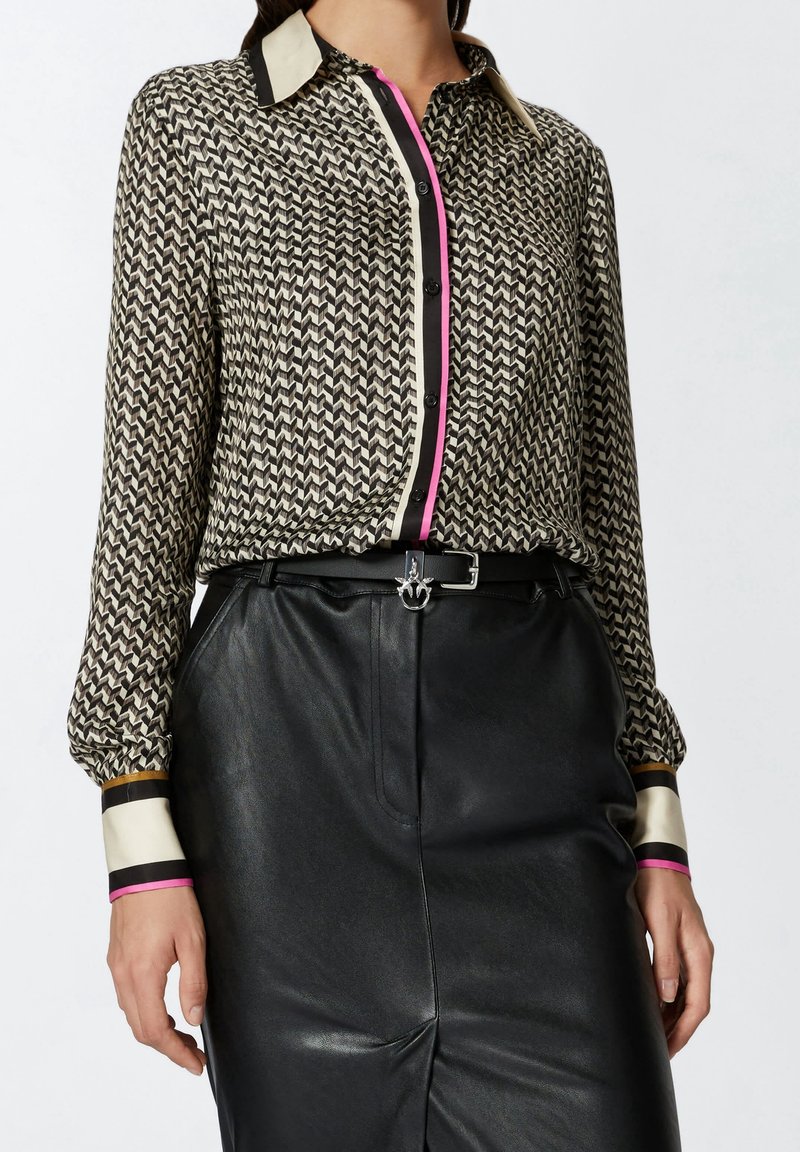 Femme portant un chemisier à manches longues à motifs noir et blanc avec des touches roses, rentré dans une jupe en cuir noire taille haute avec ceinture.