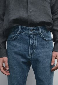 Mörkgrå skjorta med knappar i kombination med blå denimjeans. Jeansen har framfickor och en lätt blekning, vilket visar en klassisk passform och stil.