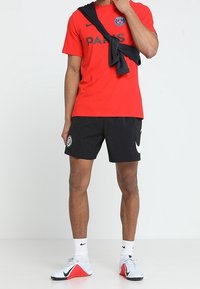 Mand iført rød PSG fodboldtrøje, sorte shorts, hvide Nike sneakers, med en sort sweatshirt bundet om skuldrene, stående på hvidt gulv.