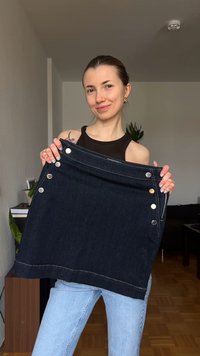 Νεαρή γυναίκα που χαμογελάει σε εσωτερικό χώρο κρατώντας σκούρα τζιν φούστα με ασημένια κουμπιά, φοράει μαύρο τοπ χωρίς μανίκια και ανοιχτό μπλε τζιν.